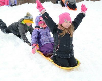 Annabelle Quast-Clark, a first grader, joyfully raises her arms for the journey down the hill at Bonner Elementary School on Monday, Jan. 20. First grader Landon Rogers pushes the sled also occupied by Payton Braswell, center, another first grader. In below photo, Julie Manley, a paraprofessional, assists first grader Brinley Palumbo down the hill.